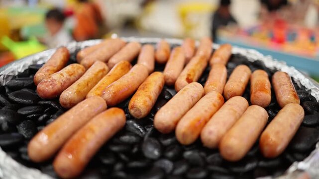 Grilled sausage on hot black stone at a food court in a supermarket. Street food cooking process in a store. Delicious roasted meat snack on a grill with blurred background concept.