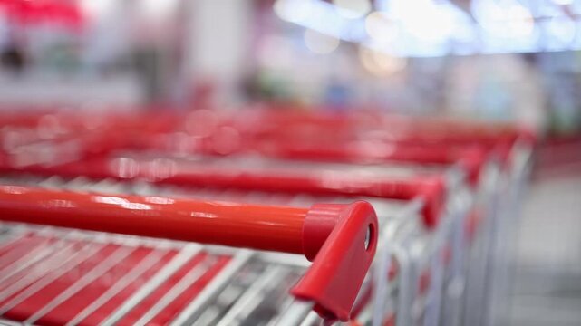 Row of red handles of shopping carts in a supermarket. Blurred store interior on background. Retail equipment for customer and consumerism concept. Empty trolley in a shop.