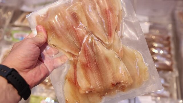Close up of a hand holding a vacuum pack of dry small fish fillet in a supermarket. Customer choosing snack or seafood product in a store. Salty dried fish in plastic package concept.
