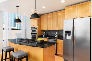 Fototapeta na wymiar A contemporary kitchen features light wood cabinets, black granite countertops, a black subway tile backsplash, and a large island with bar stools. No brands or labels.