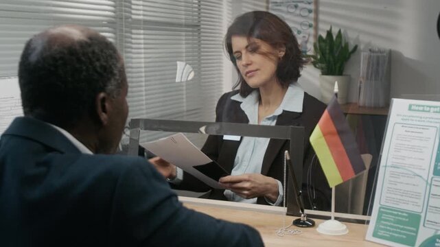 Rear medium close-up of senior African American businessman applying for travel visa at German consulate, handing application form, passport and photos to Caucasian female service agent