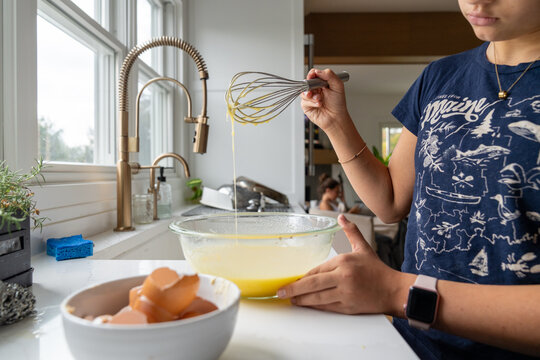 Teenage girl whisking eggs at kitchen sink in bright modern kitchen