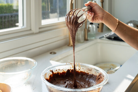 Chocolate brownie batter dripping from whisk in sunlit kitchen window