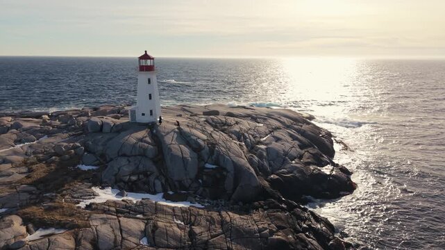 Cinematic aerial sunset view: Peggy's Cove iconic lighthouse on snow-blanketed rocks by Atlantic. Halifax Nova Scotia Canada winter scene, golden hour beauty 4K.