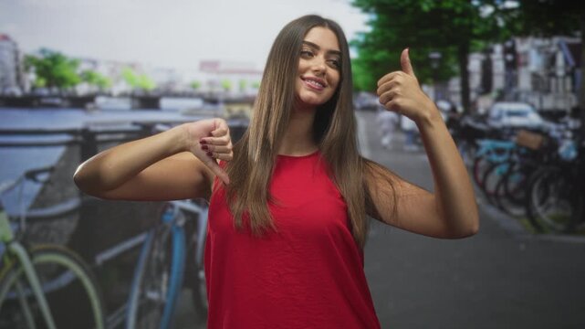 Young hispanic woman shows thumbs up with right hand and thumbs down with left, wearing red top on street; ambivalence.
