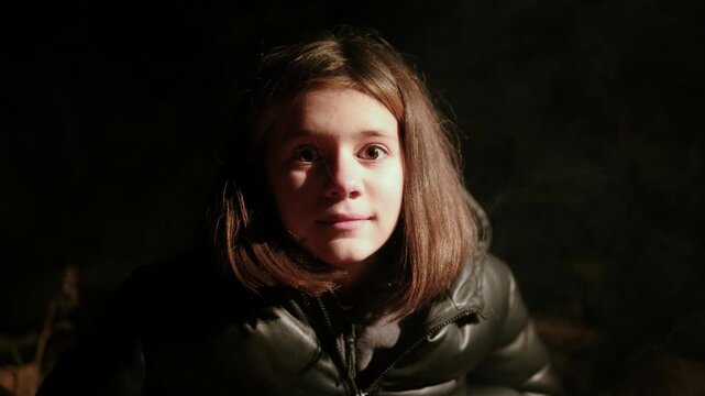 Close-up portrait of a young girl with an expressive face illuminated by campfire light, listening intently to a spooky story in the dark, with smoke wafting in the background at night