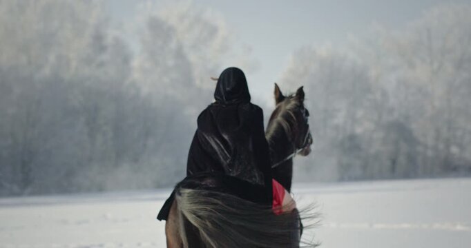 Hooded cloaked figure riding horse in winter snow
