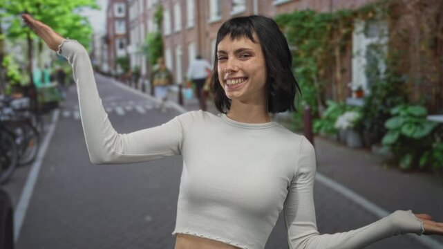 Young hispanic woman with bare midriff raising arms and smiling on narrow street in amsterdam; confidence.