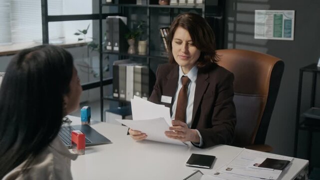 Medium shot of middle-aged Hispanic woman arriving for interview with Caucasian female consular officer, who is returning incomplete visa application form and explaining requirements