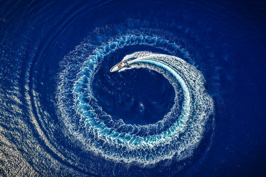 High aerial overhead view of a motor RIB boat forming a circle of bubbles and surf with its engine over blue ocean