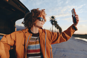 Fototapeta premium Smiling woman with curly hair wearing rainbow sweater and orange jacket taking selfie with smartphone near car outdoors. Lifestyle concept with casual clothing and sunny weather.