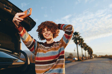 Fototapeta premium Smiling woman with curly hair wearing a rainbow sweater takes a selfie with a smartphone near a car on a sunny day. Outdoor lifestyle with palm trees and clear sky background.