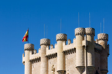 Close view of the tower section of a medieval castle in Segovia, Spain, with decorative stone turrets and the Spanish flag against a clear blue sky. © Alexey Bakharev