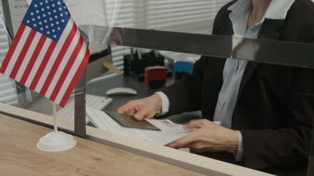 Close-up of hands of unrecognizable female USA consular officer putting red rejection stamp on visa application, then returning to customer while working behind glass partition at embassy