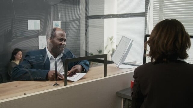 Medium shot of smiling middle-aged African American businessman approaching counter with glass partition at visa center, greeting female officer and submitting set of documents for work visa