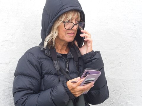 A lady with hood up speaks on a smartphone and holds another. mobile phone in other hand. She has a worried concerned look on her face  and has binoculars around her neck	