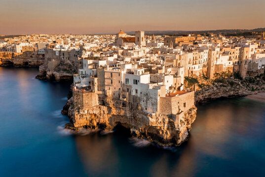 Aerial view of the historic white buildings perched on limestone cliffs overlooking the turquoise sea during sunset in Polignano a Mare, Apulia, Italy.