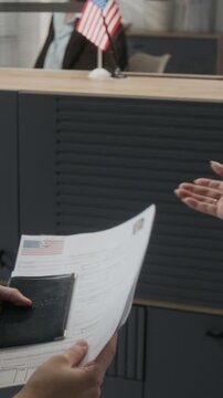 Over-shoulder vertical shot of anonymous woman and female friend checking documents, then submitting to USA embassy clerk while applying for American immigration visa