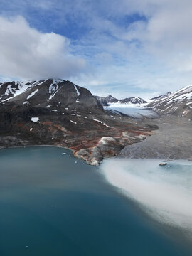 Aerial view of rugged snow-capped mountains and a glacier flowing into a turquoise sea under a cloudy sky in Greenland.