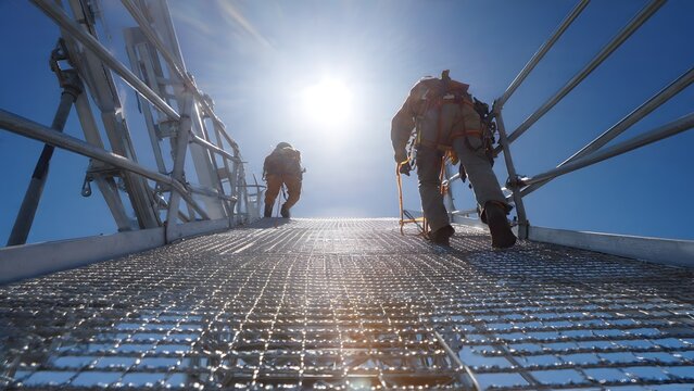 Two high altitude workers climbing a steel mesh walkway atop a roof structure, both tied to overhead fall arrest rails, sun reflecting off metal