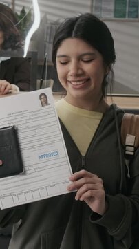Medium vertical portrait of cheerful young Hispanic girl posing at consulate after interview, holding paperwork and passport, happy at student visa approval, looking at camera, smiling with joy