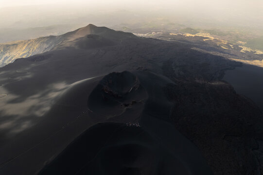 Aerial view of the volcanic craters and dark lava fields of Mount Etna with a winding path under a hazy sky in Castiglione di Sicilia, Sicily, Italy.