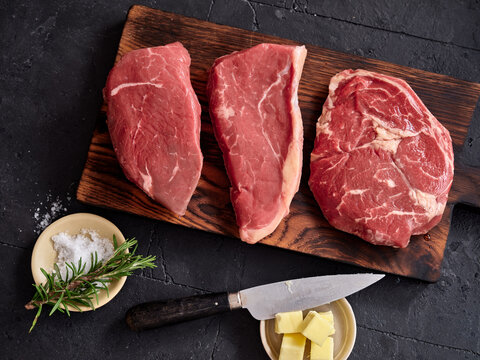 Raw rib-eye, roast beef, flank and rump steak on a wooden board with knife and butter at a kitchen counter