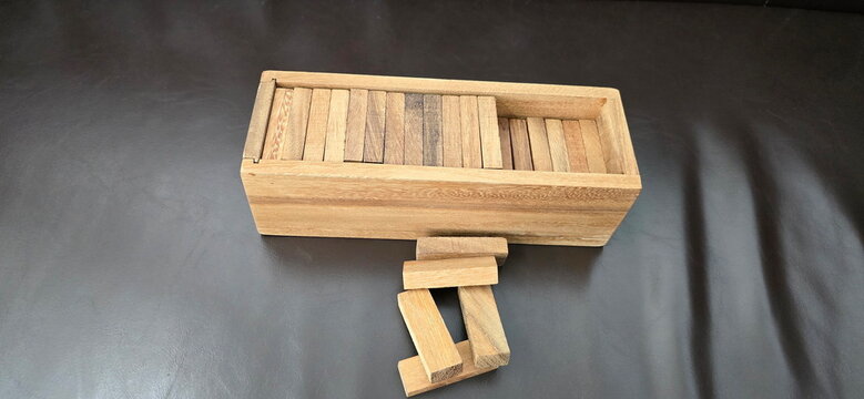 The box contained rectangular wooden blocks for game Jenga. However, some blocks were missing from box, some were piled outside the box in preparation for building tower. Game on a brown background.
