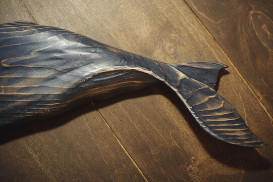 Horizontal close-up of a handcrafted wooden sperm whale tail with a deep burnt texture, showcasing the intricate carved fins against a dark wood plank background.