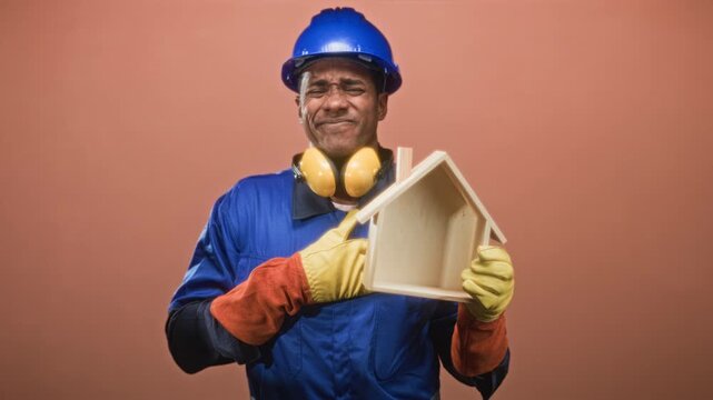 Man builder in hardhat pointing finger into small wooden house model while holding it with gloved hands in studio; pride.
