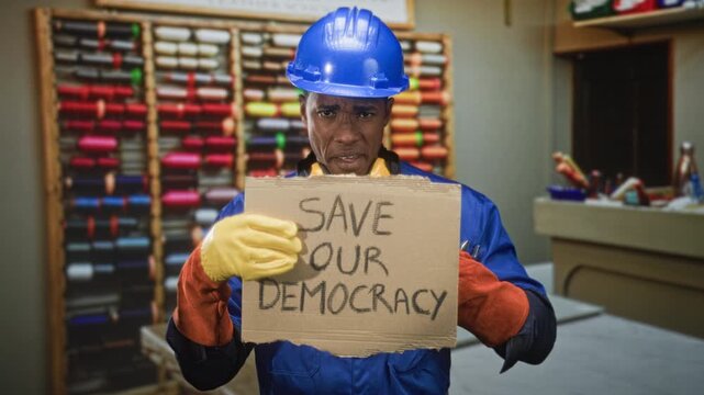 Man in hardhat and coveralls holding a cardboard sign reading save our democracy in a workshop with spools and tools visible; worker solidarity determination.