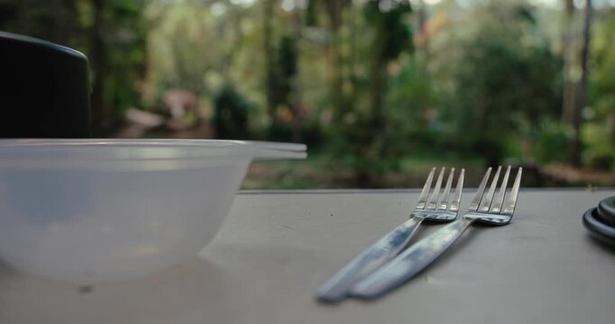 Metal forks and plastic bowl resting on outdoor table. Cutlery and dishware set for meal in garden or forest environment with natural lighting and blurred background.
