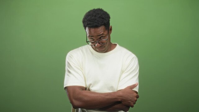 Man adjusts glasses with finger, arms crossed in studio before green backdrop, wearing white tee; skeptical dry humor.