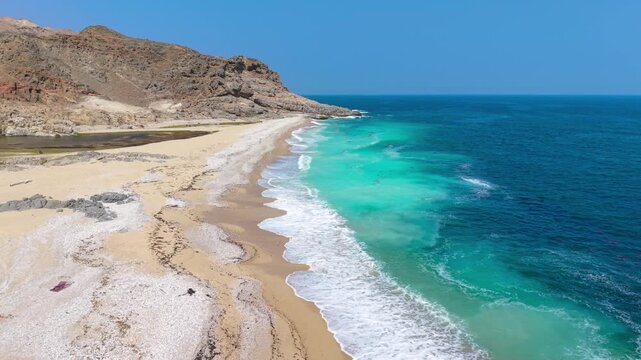 Aerial view of turquoise water and waves crashing onto a sandy beach near rocky cliffs under a clear blue sky, Saudi Arabia.