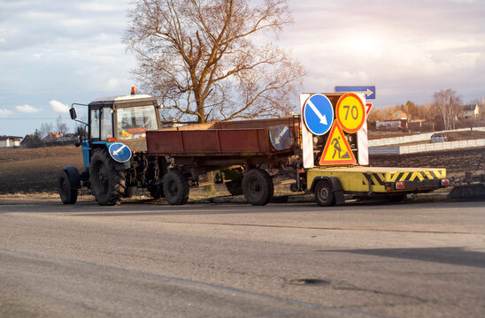Blue agricultural tractor transporting two trailers, one empty, other carrying bright yellow road construction warning signs, on side of asphalt road near rural field.