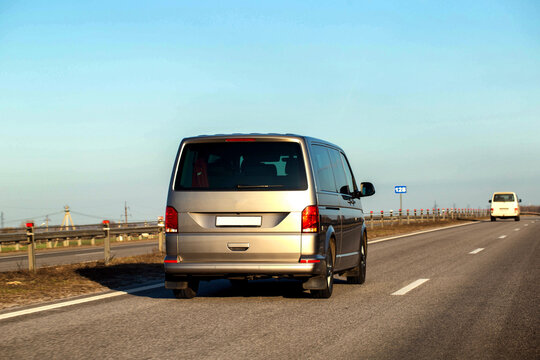 Rear view of silver minivan traveling on asphalt highway under clear blue sky, guardrail along road edge, other vehicles in distance