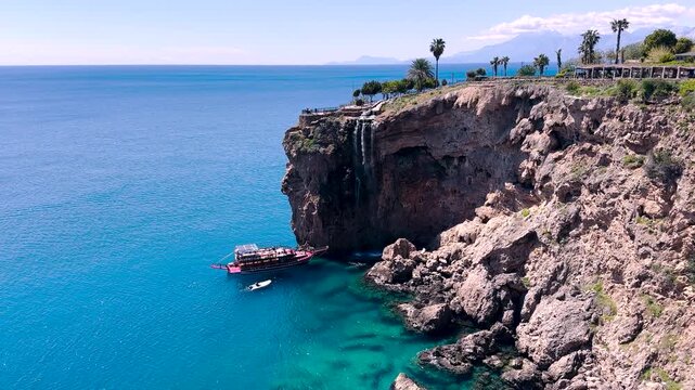 Scenic Antalya T&uuml;rkiye coast with dramatic cliffs and a small waterfall flowing into the Mediterranean. A tour boat and speedboat move across turquoise water under a clear blue sky.