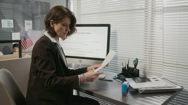 Medium side shot of mature Caucasian female USA consulate official putting approval stamp on American business or travel visa application, customers queuing for appointment in background