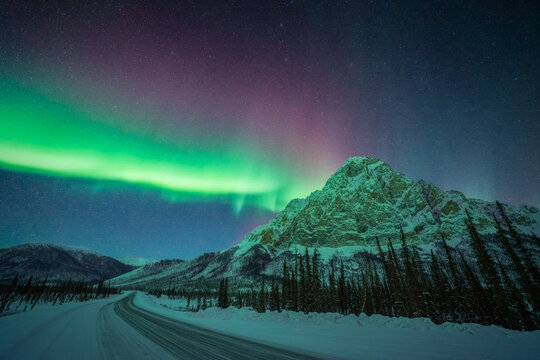 View of the Aurora Borealis dances above the snow-covered road and majestic, snow-capped mountain peaks under a starlit sky, Coldfoot, Alaska, United States.