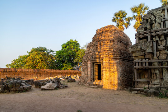 View of ancient stone structures glowing warmly under a soft light, with trees and a long wall adding depth, creating a serene scene, Siem Reap, Siem Reap Province, Cambodia.