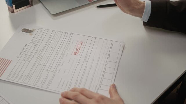 Close-up of hands of anonymous American female consular officer putting rejected stamp on USA work visa application for black man, after assessment and verification procedure