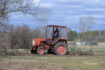 Rolnik w czerwonym ciągniku orze, sprężynuje glebę, ziemię w ogródku przydomowym na wsi © Joanna