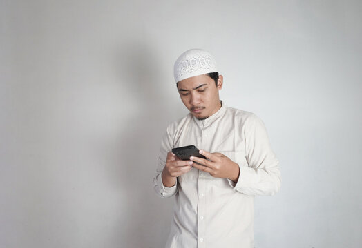 Asian Muslim man is busy with his smartphone. Man with cream colored koko shirt and white peci. Copy space. Promotion Concept for Islamic Market