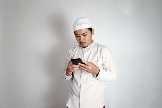 Asian Muslim man is busy with his smartphone. Man with cream colored koko shirt and white peci. Copy space. Promotion Concept for Islamic Market