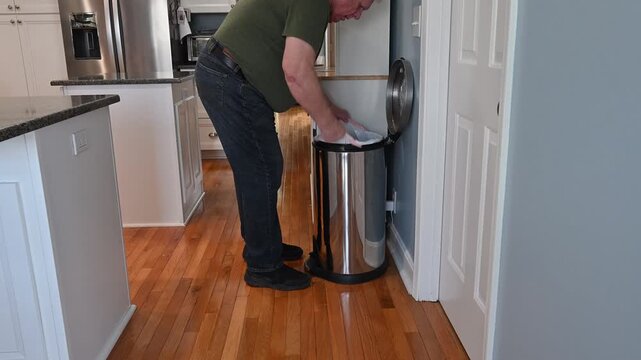 Older white man removes trash bag from stainless steel bin in modern kitchen