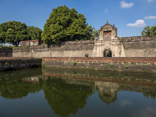 Obraz premium Historic Puerta Real gate of Intramuros in Manila, Philippines. Ancient stone walls and ornate archway reflected perfectly in the serene moat, showcasing colonial architectural grandeur under a bright