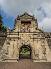Obraz premium The majestic entrance to Fort Santiago in Intramuros, Manila, Philippines, revealing historic stone architecture, intricate bas-reliefs, and a grand archway under a dramatic cloudy backdrop.