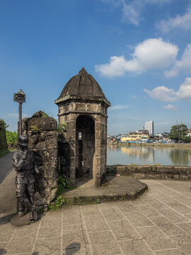 Historic stone garita and bronze soldier statue at Fort Santiago, Intramuros, Manila, Philippines. Overlooking a waterway with a bustling city skyline in the distance on a clear day.