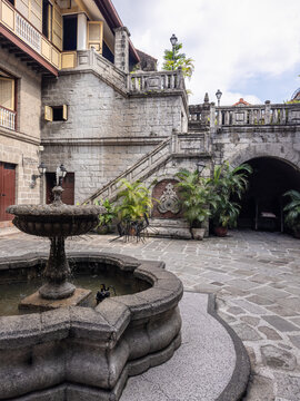 Historic stone courtyard at Las Casas Filipinas de Acuzar, Bataan, featuring a charming fountain, traditional colonial architecture, winding staircases, and lush potted plants under a bright sky.
