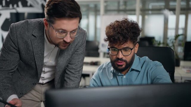 Two multiethnic business men looking in computer monitor cooperate working online job Caucasian leader with digital tablet help teaching advice to Arabian intern diversity office businessmen talking